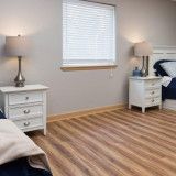 Bedroom with two beds, two white nightstands with lamps, a window with blinds, and wood-look flooring.