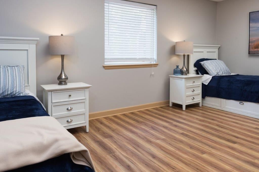 Bedroom with two beds, white nightstands, lamps, window with blinds, and wood-look flooring.