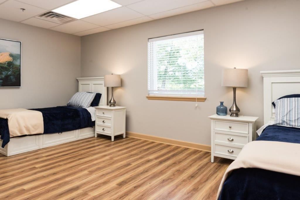 Two beds in a room with nightstands, lamps, and a window. Light wood floor and gray walls.