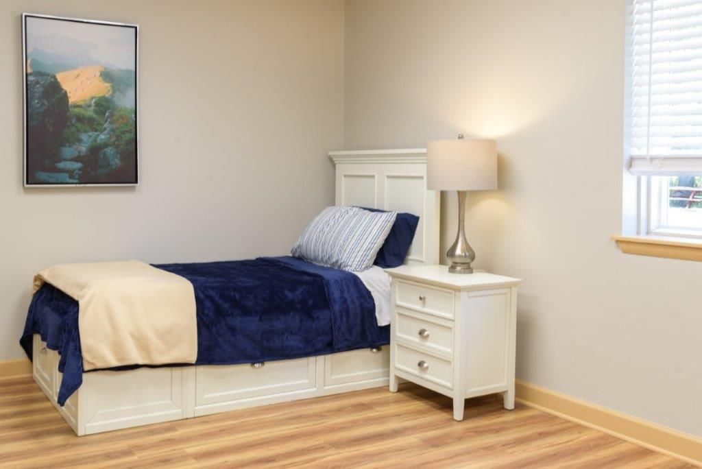 Bedroom with a white bed, nightstand, and window. Artwork hangs on the wall.