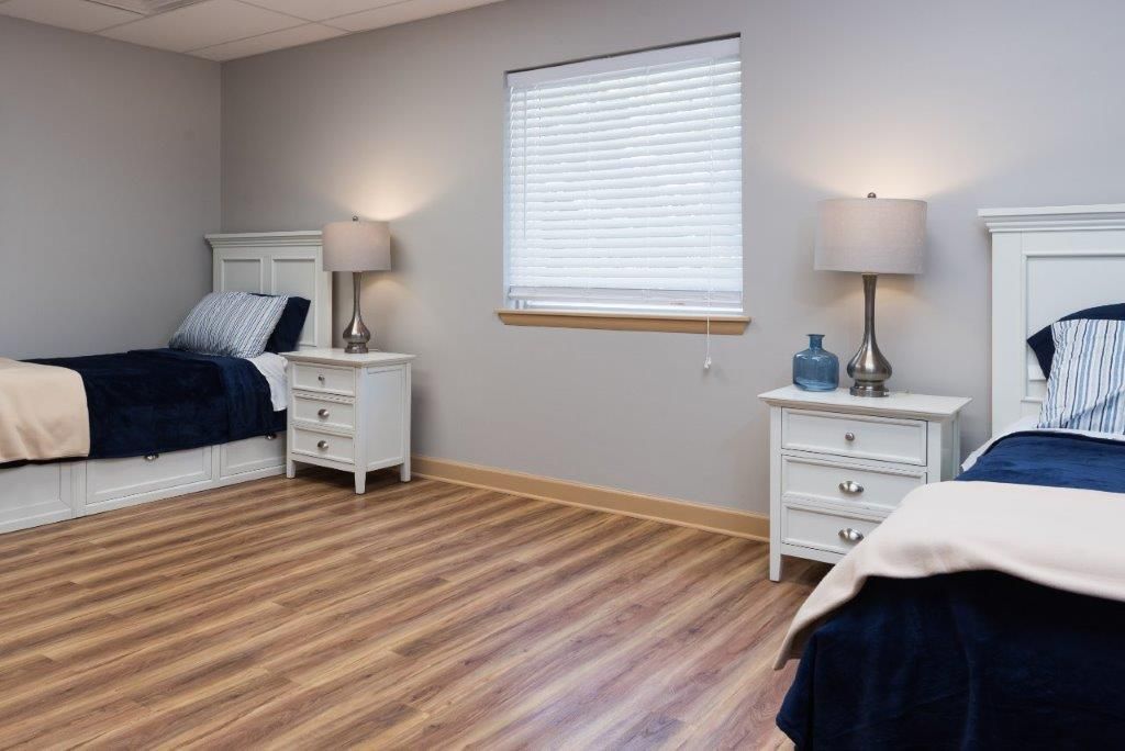 Two twin beds in a room with white nightstands, lamps, and a window with blinds.