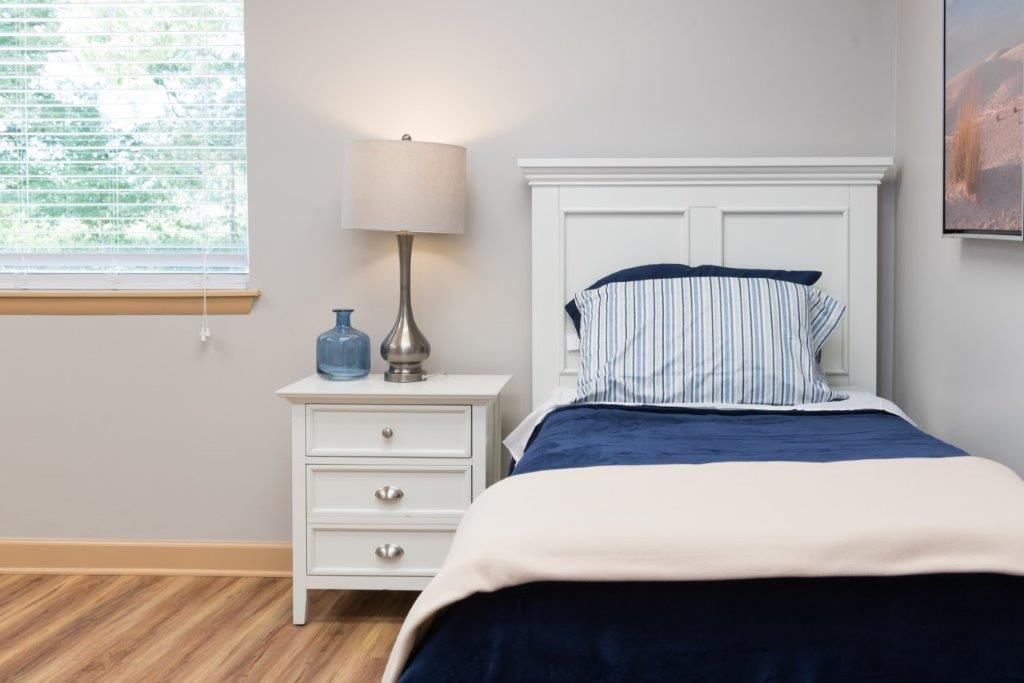 White bedroom with a single bed, nightstand, lamp, and window. Blue and white bedding.