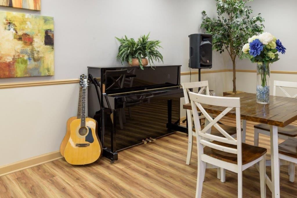 Room with a piano, guitar, table, chairs, and flowers. Black piano next to a guitar, table with flowers.