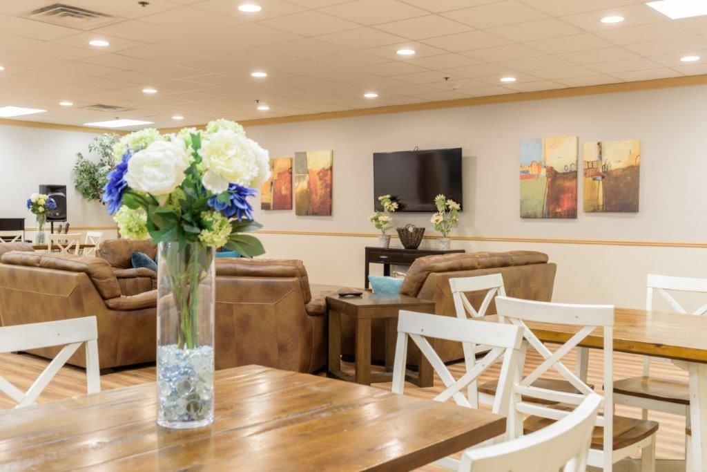 Lounge area with brown sofas, tables, white chairs, floral arrangement, TV, and artwork on the wall.