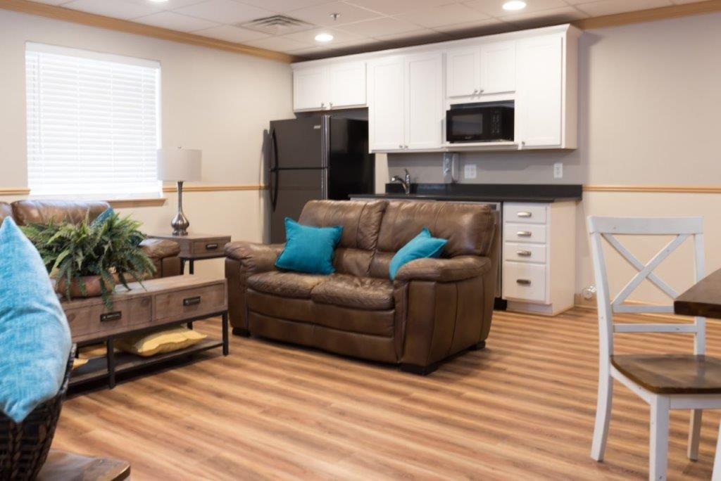 Small living space with kitchen area; brown leather sofa, white cabinets, black fridge, and laminate flooring.