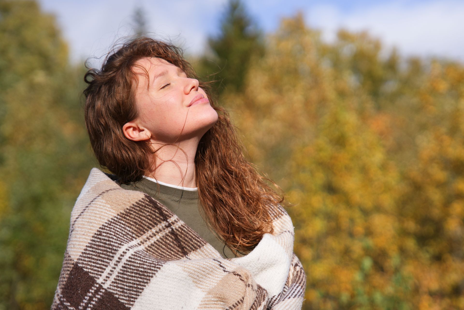 Woman wrapped in plaid blanket, eyes closed, face tilted to sunlight; autumn foliage background.