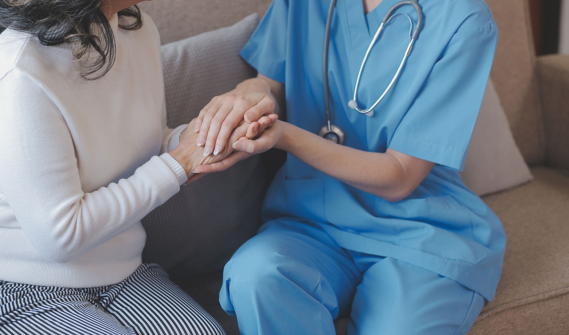 Healthcare worker in blue scrubs holding hands with a seated patient on a couch