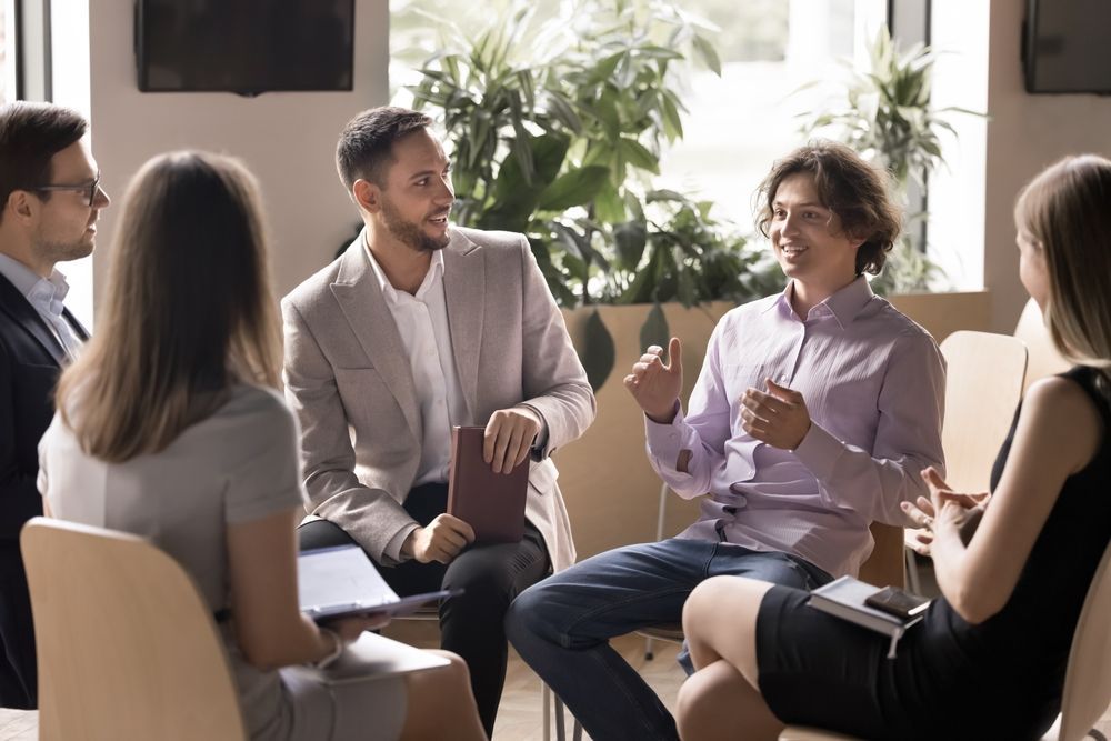 People seated in a circle, engaged in a discussion; bright, modern office with plants.