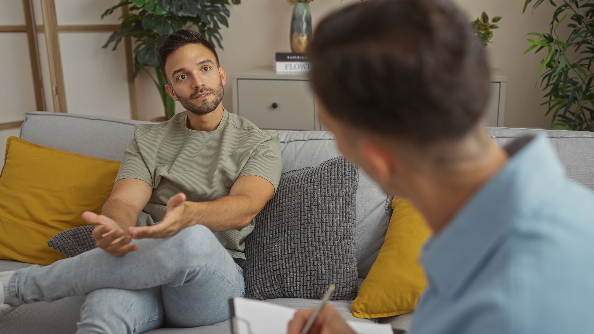 Man talking, gesturing, on couch; another person facing him, taking notes.