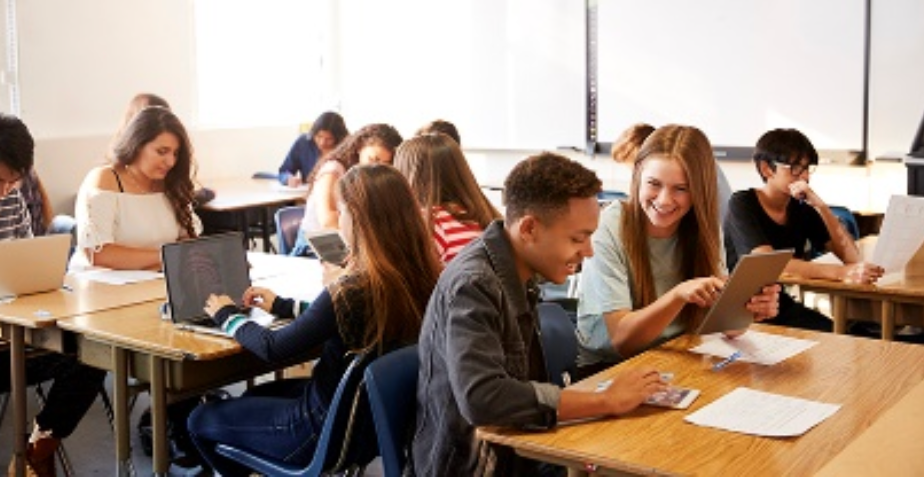 A group of students are sitting at desks in a classroom using laptops and tablets.