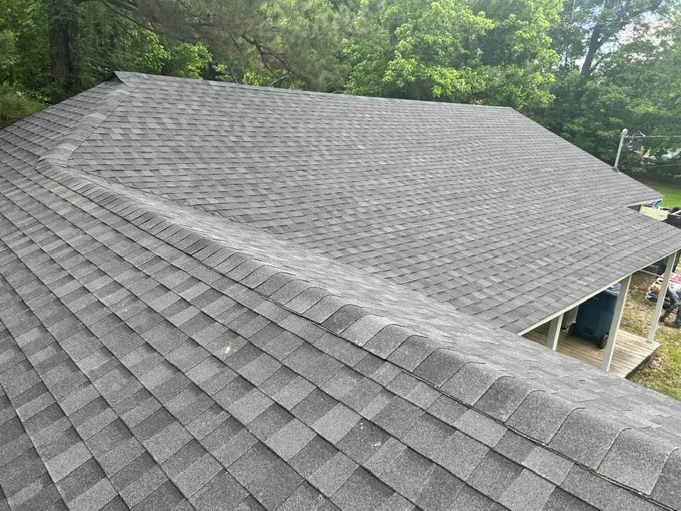 Gray asphalt shingle roof on a house, viewed from a high angle, with a backdrop of green trees.