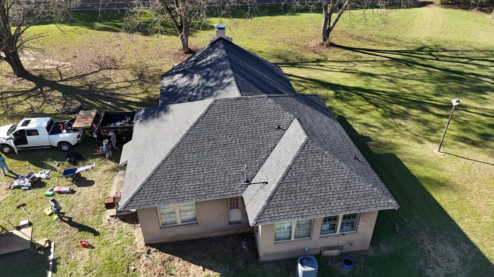 Aerial view of a house with a dark roof, surrounded by grass and trees. A white truck is parked nearby.