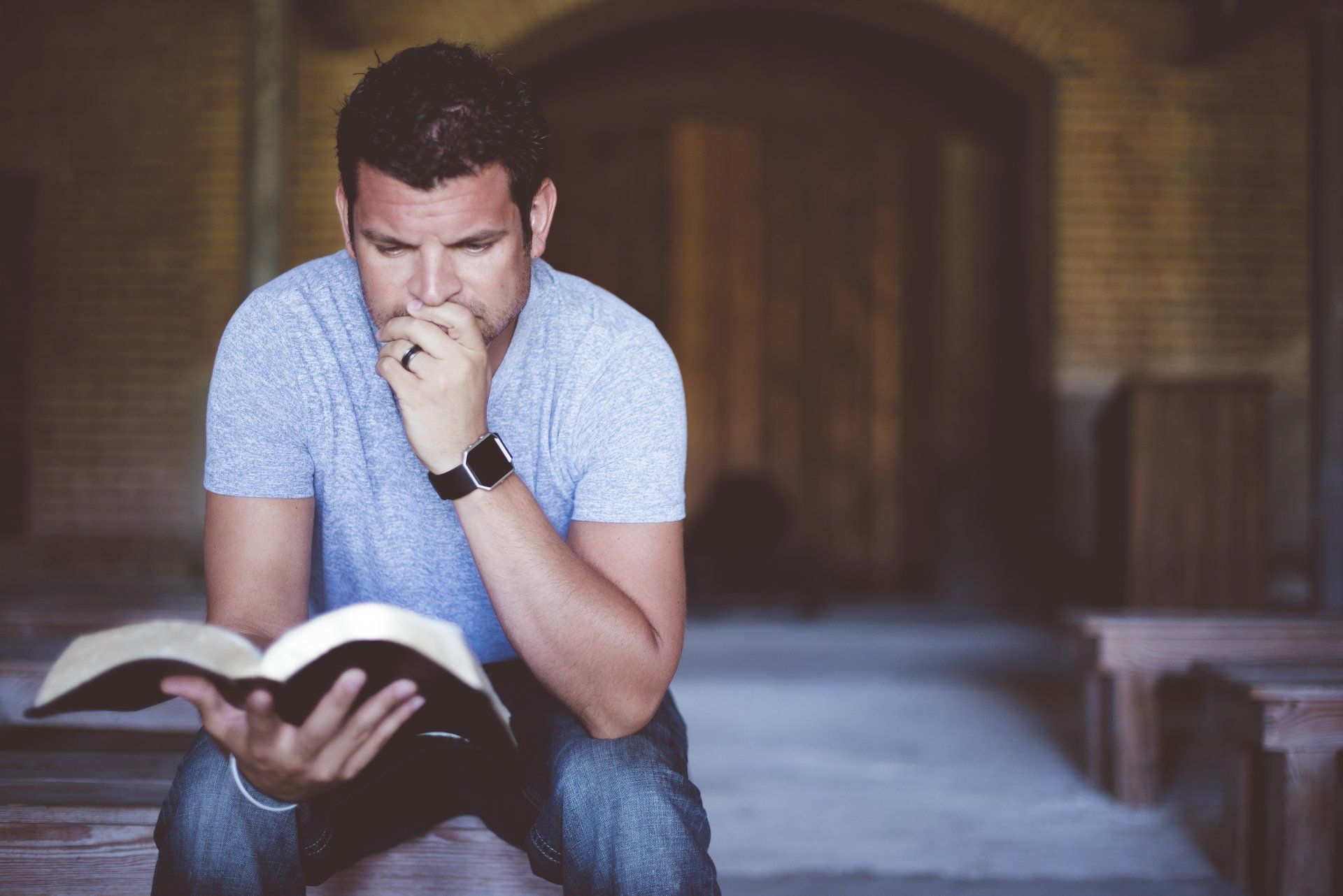 A person sitting in a rustic, dim interior, resting their chin on their hand while reading an open book.