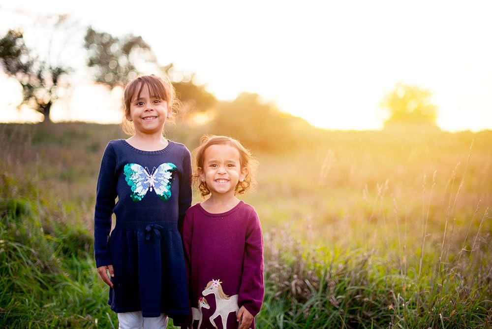 Two children smiling in a sunlit grassy field, wearing long-sleeved dresses with animal patterns.