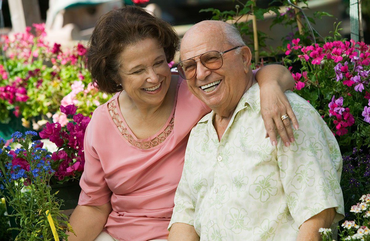 An older couple embraces and smiles while surrounded by a colorful outdoor flower garden.