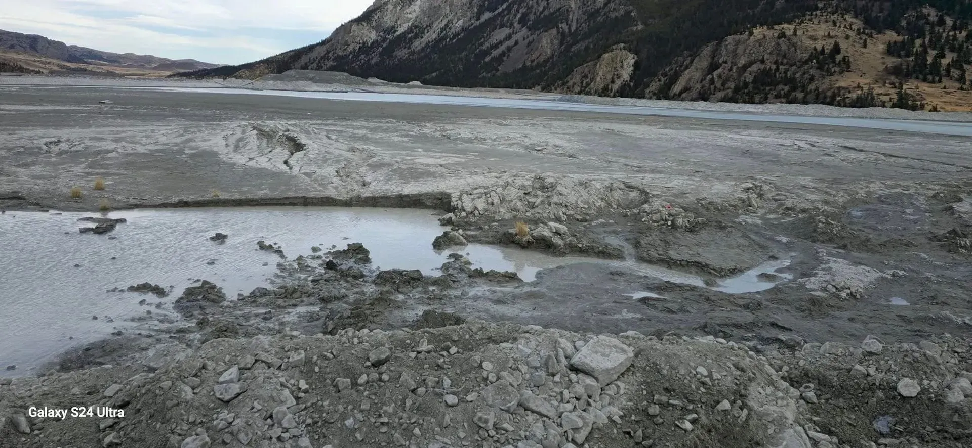 Muddy, gray terrain with water pools, mountains in the background. Cloudy sky.