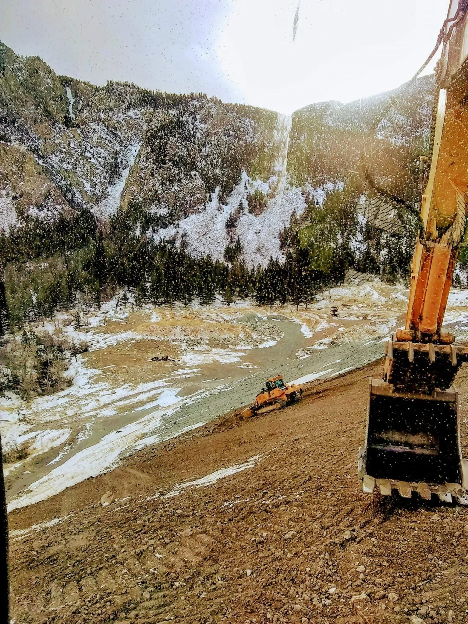 An excavator works on a snowy mountain riverbed, with mountains in the background.