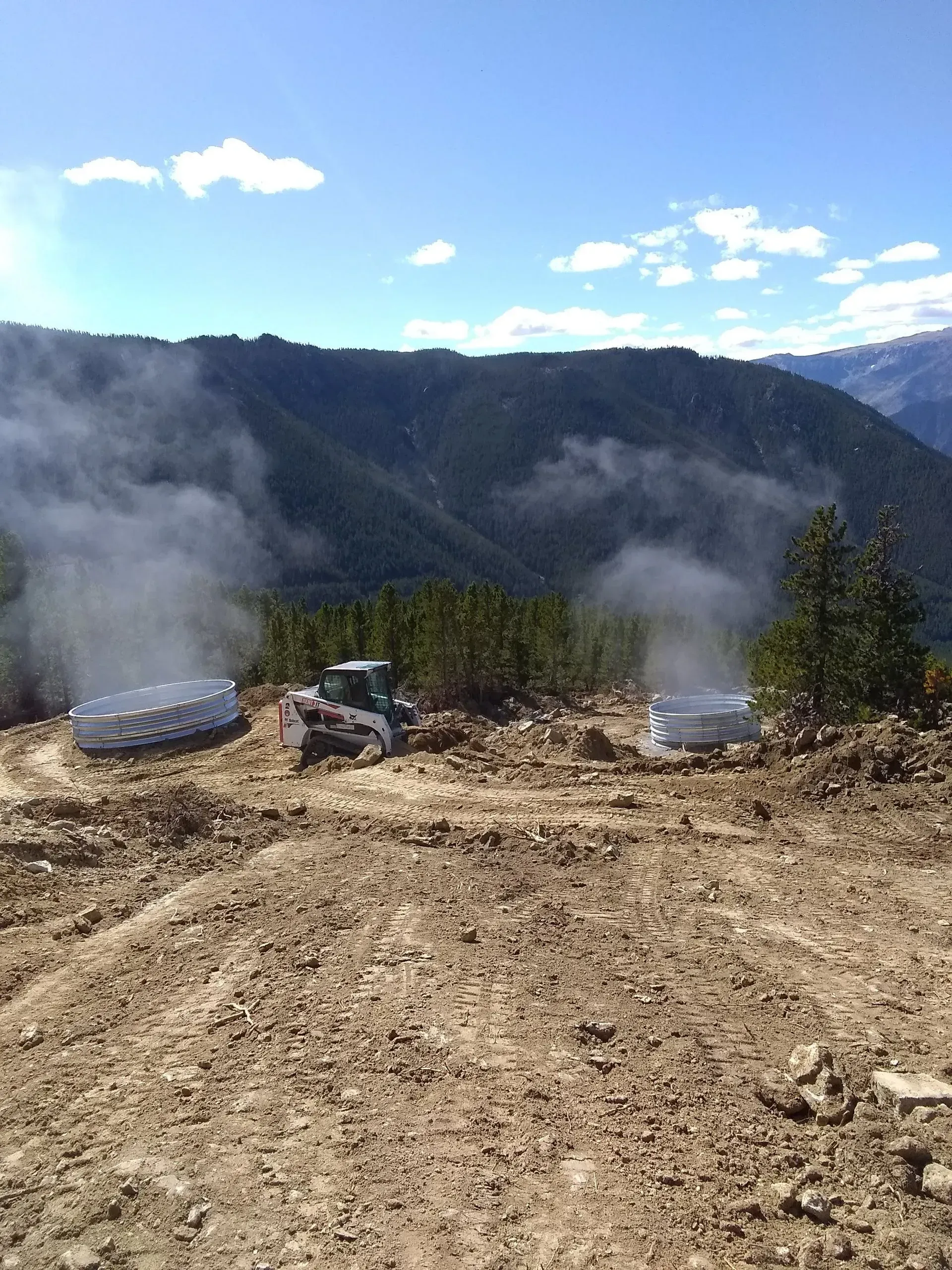 A bulldozer works on a dirt clearing, with two smoking containers, forest and mountains in the background.