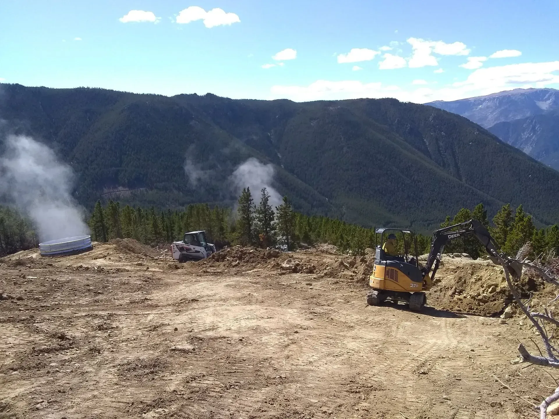 Construction site on a mountain with heavy machinery. Dust and trees in the background.