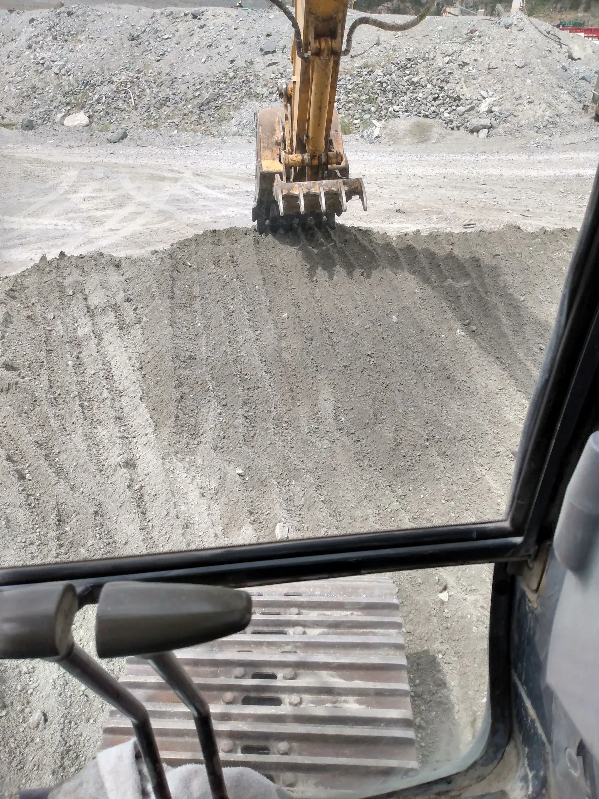View from inside an excavator cab, overlooking gray gravel pile being moved by the excavator's arm in a quarry.