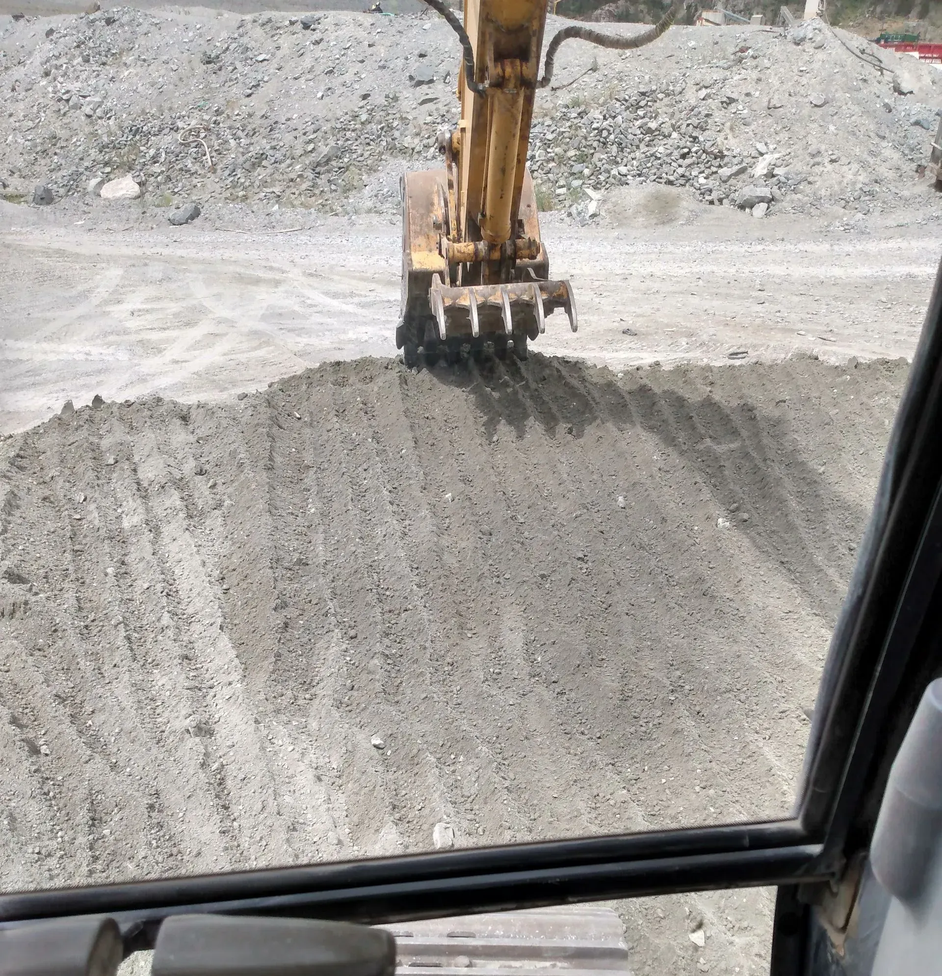 An excavator leveling a pile of gray gravel in a construction setting, viewed from inside the cab.