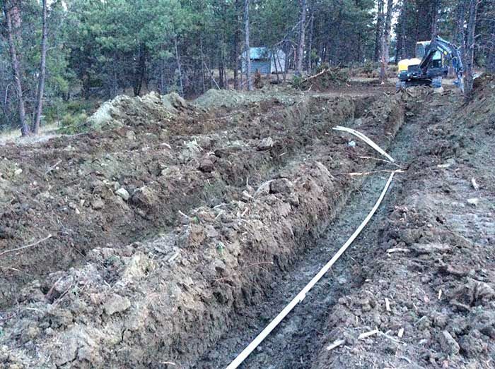 Trenches dug in earth, with a white pipe laid in one; excavator in background.