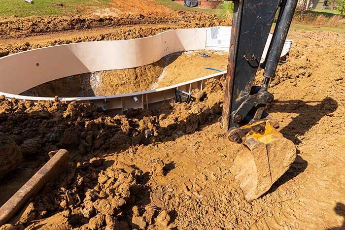 Excavator digging around a partially installed, beige-walled swimming pool in a dirt yard.