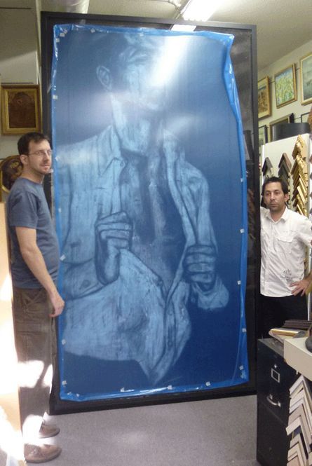 Two men holding a large framed blue artwork depicting a man in a suit. Inside a framing shop.