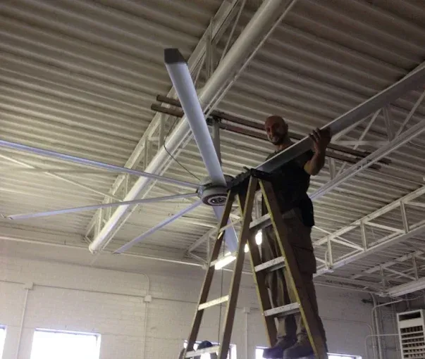 Man on ladder installing a large white ceiling fan in a building with a metal roof.