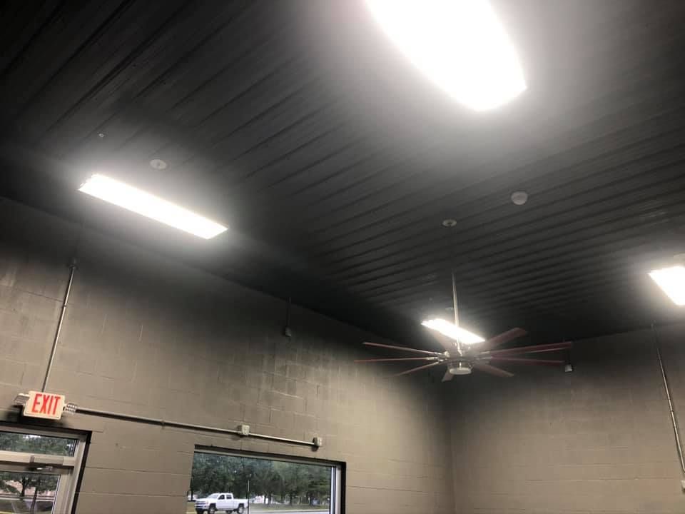 Gray interior with fluorescent lights, ceiling fan, and an exit sign. An open doorway reveals an outdoor view.