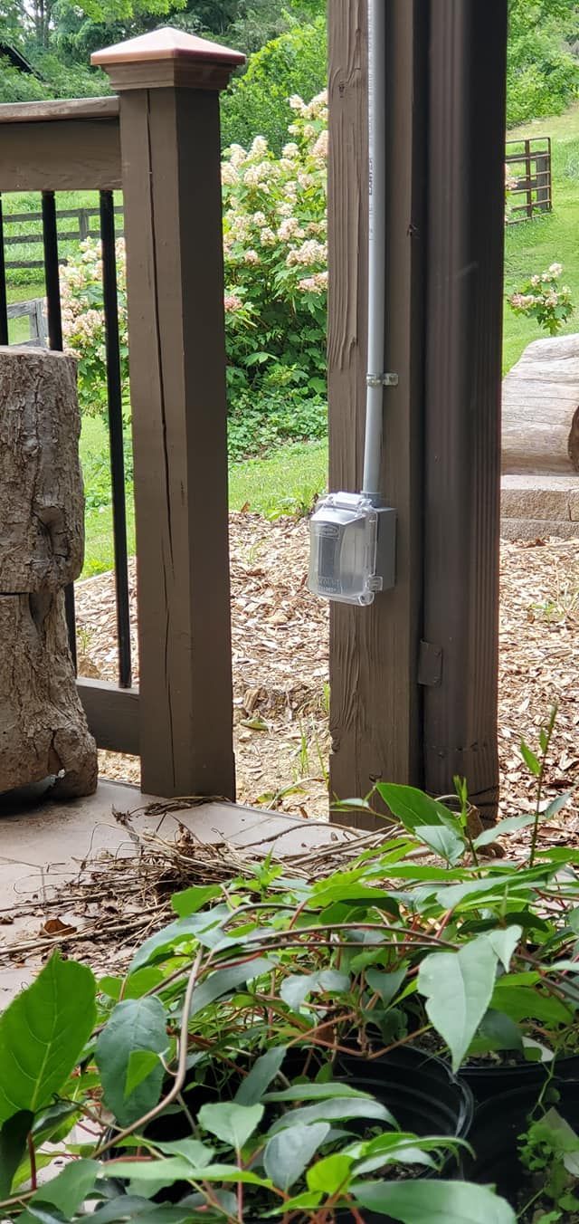 Electrical outlet box and conduit on a wooden porch post with green foliage in the foreground and a grassy yard in the background.