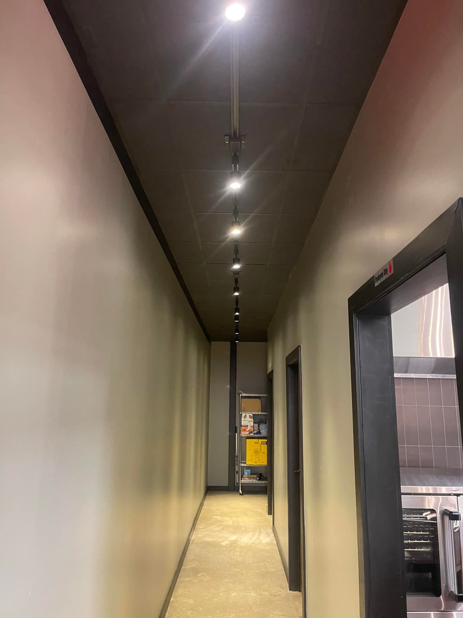 A narrow hallway with a dark ceiling and track lighting. White walls, a door on the right.