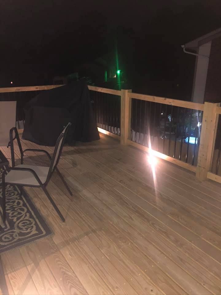 Wooden deck at night, black grill and chairs. Lit railing with dark sky.