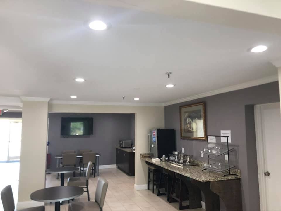Hotel breakfast area with tables, chairs, coffee station, and television. Gray and beige color scheme.