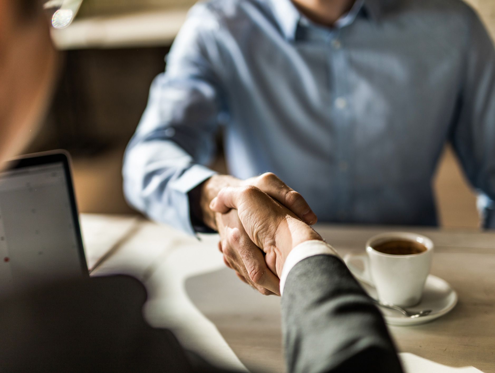 Two men are shaking hands over a table with a cup of coffee.