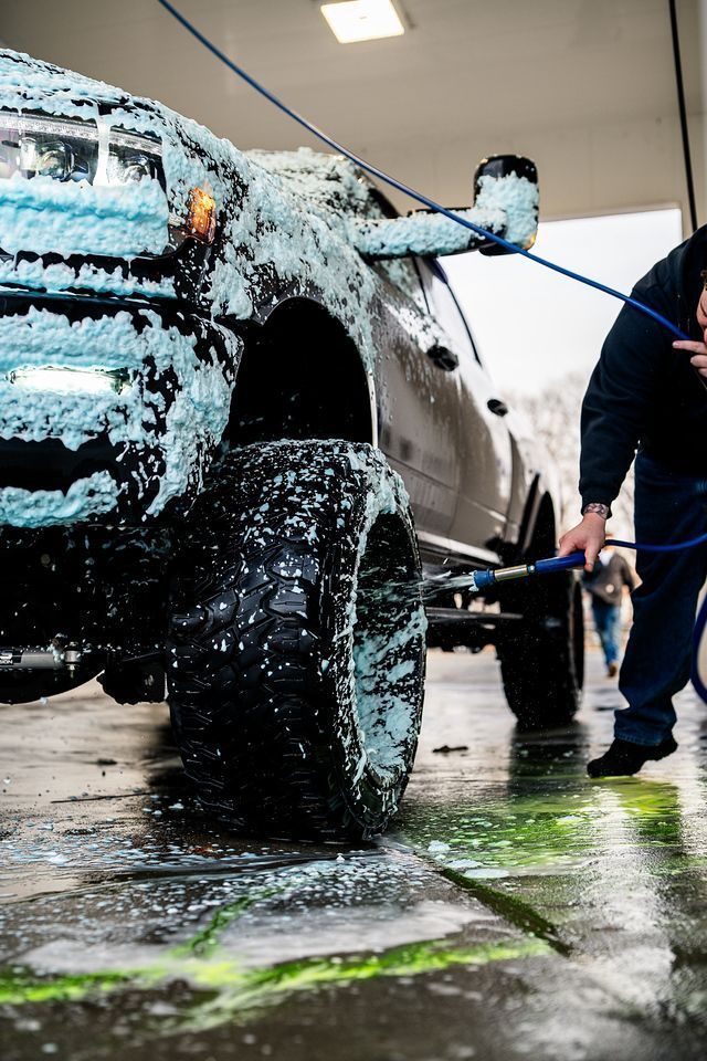 Truck being washed with foamy soap at a car wash. A person holds a hose.