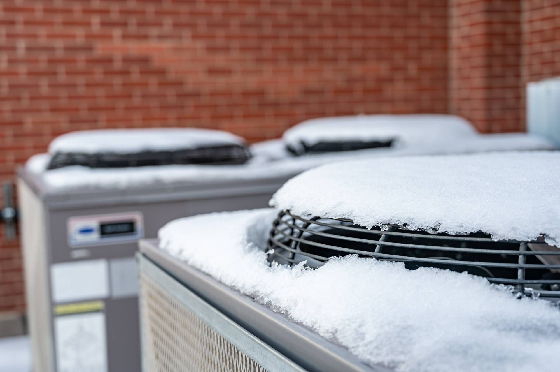 A collection of three air conditioning units outside of a building, covered in freshly fallen snow n