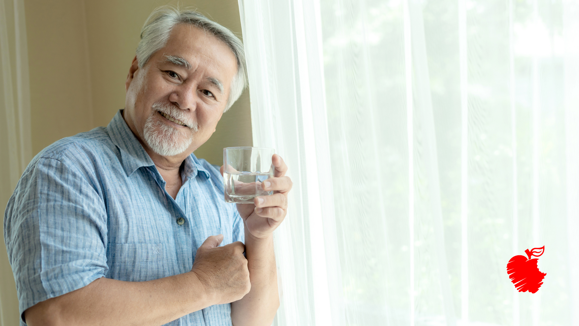 An elderly man is holding a glass of water in front of a window.