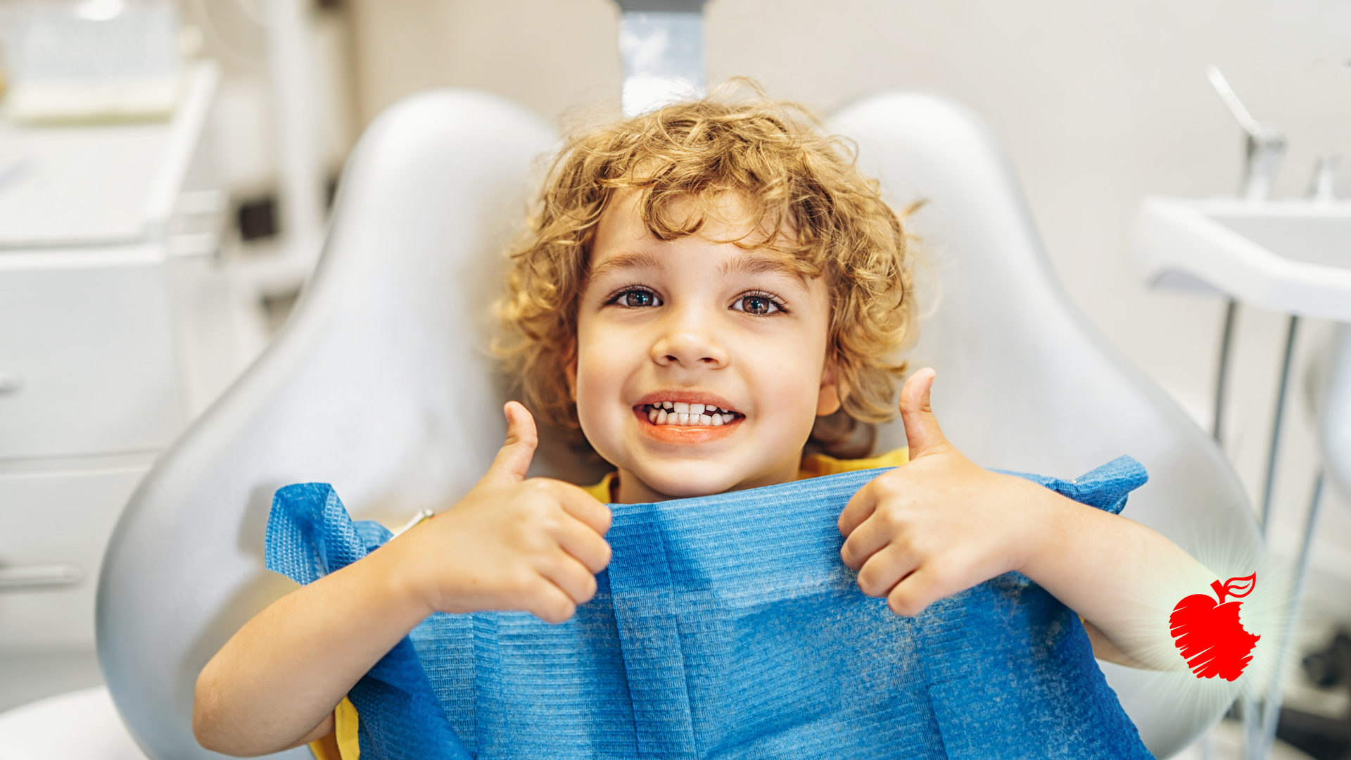 Young child with curly hair in a dentist chair giving thumbs up, smiling. Blue bib, bright setting.