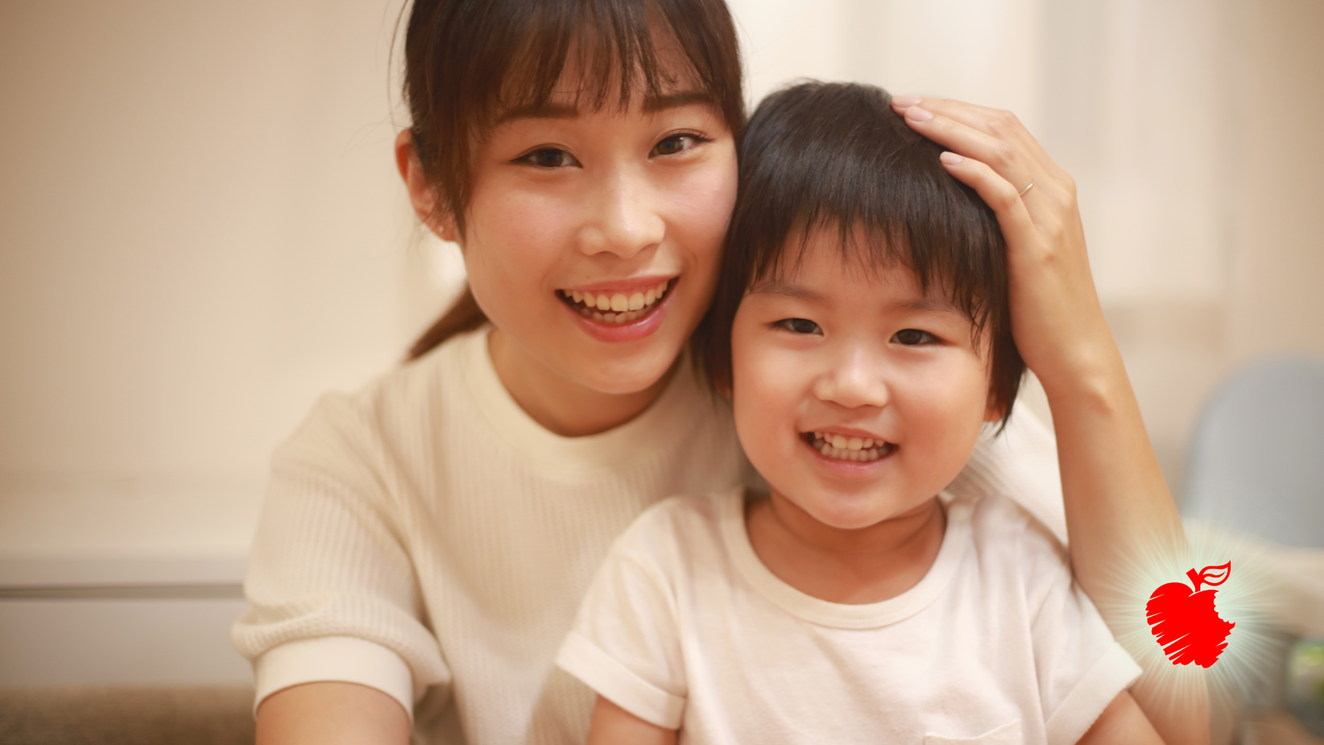 Woman and child smiling, woman's hand on the child's head, both looking at the camera. Indoor setting.