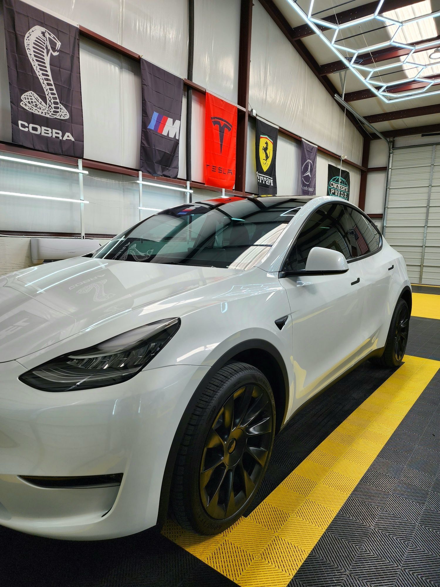 White Tesla Model Y car parked on a checkered garage floor, with car brand flags in the background.