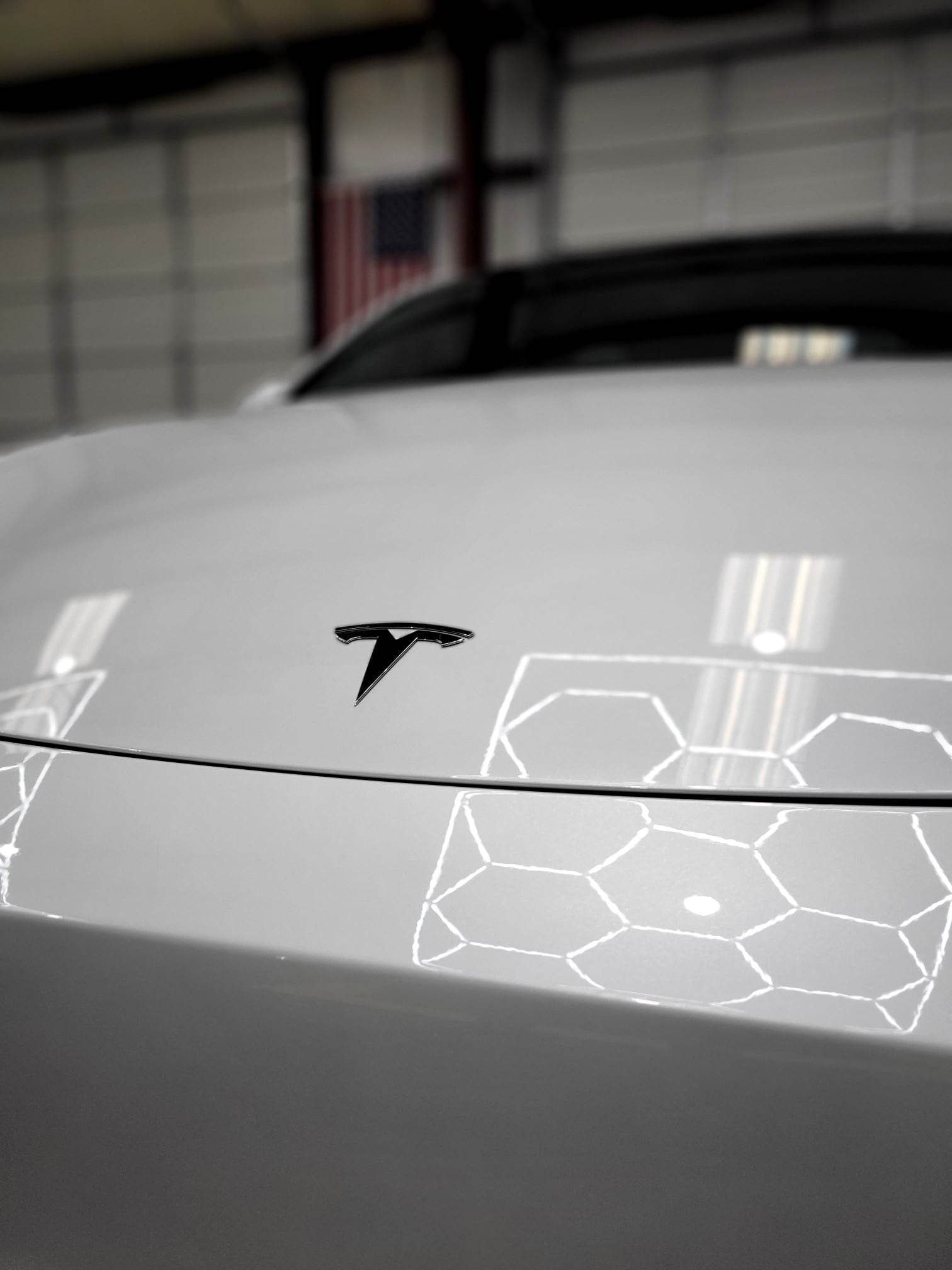 White Tesla car hood with the company logo, reflecting hexagonal patterns from overhead lights.