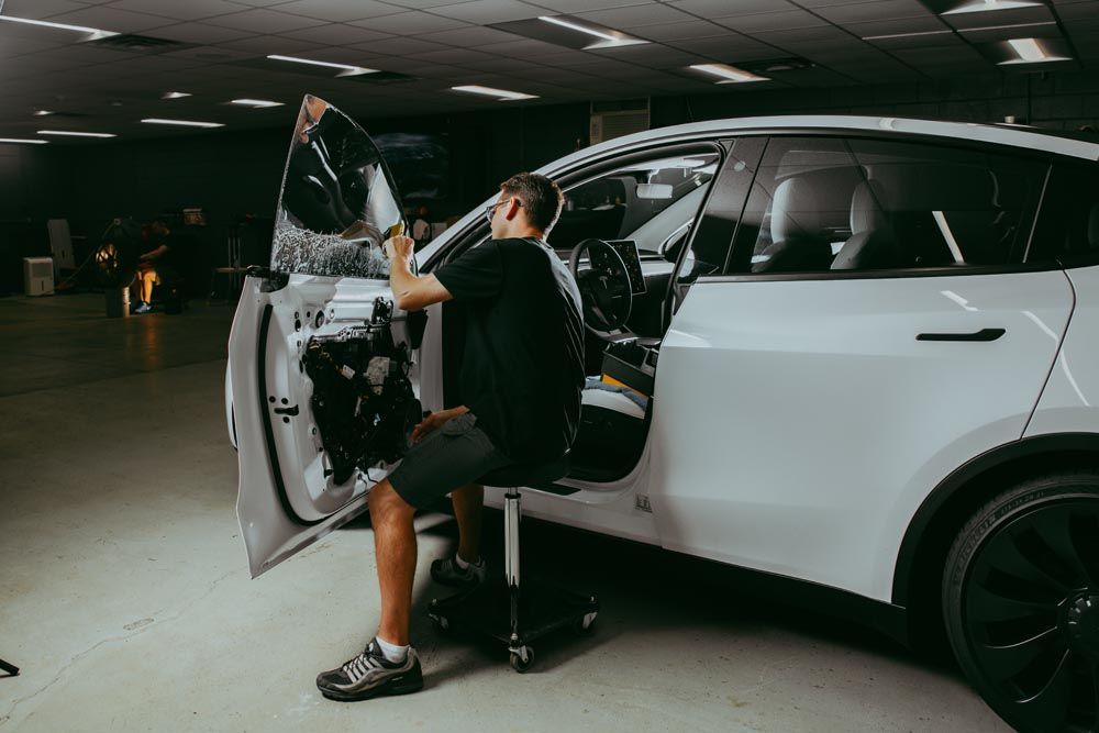Man installing tinted window film on a white car in a garage setting.