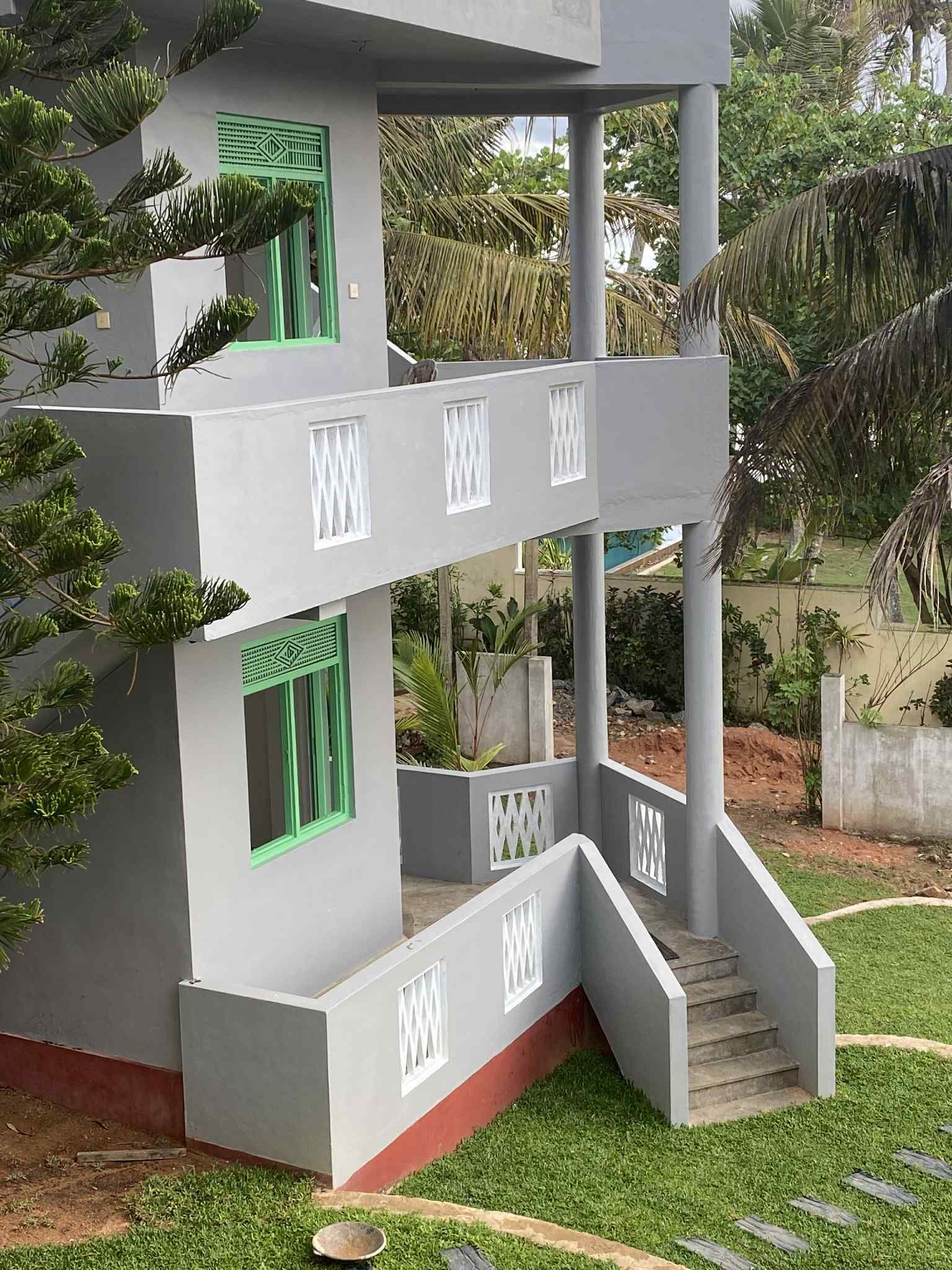Two-story gray building with white trim, green windows, and small balcony overlooking grass.
