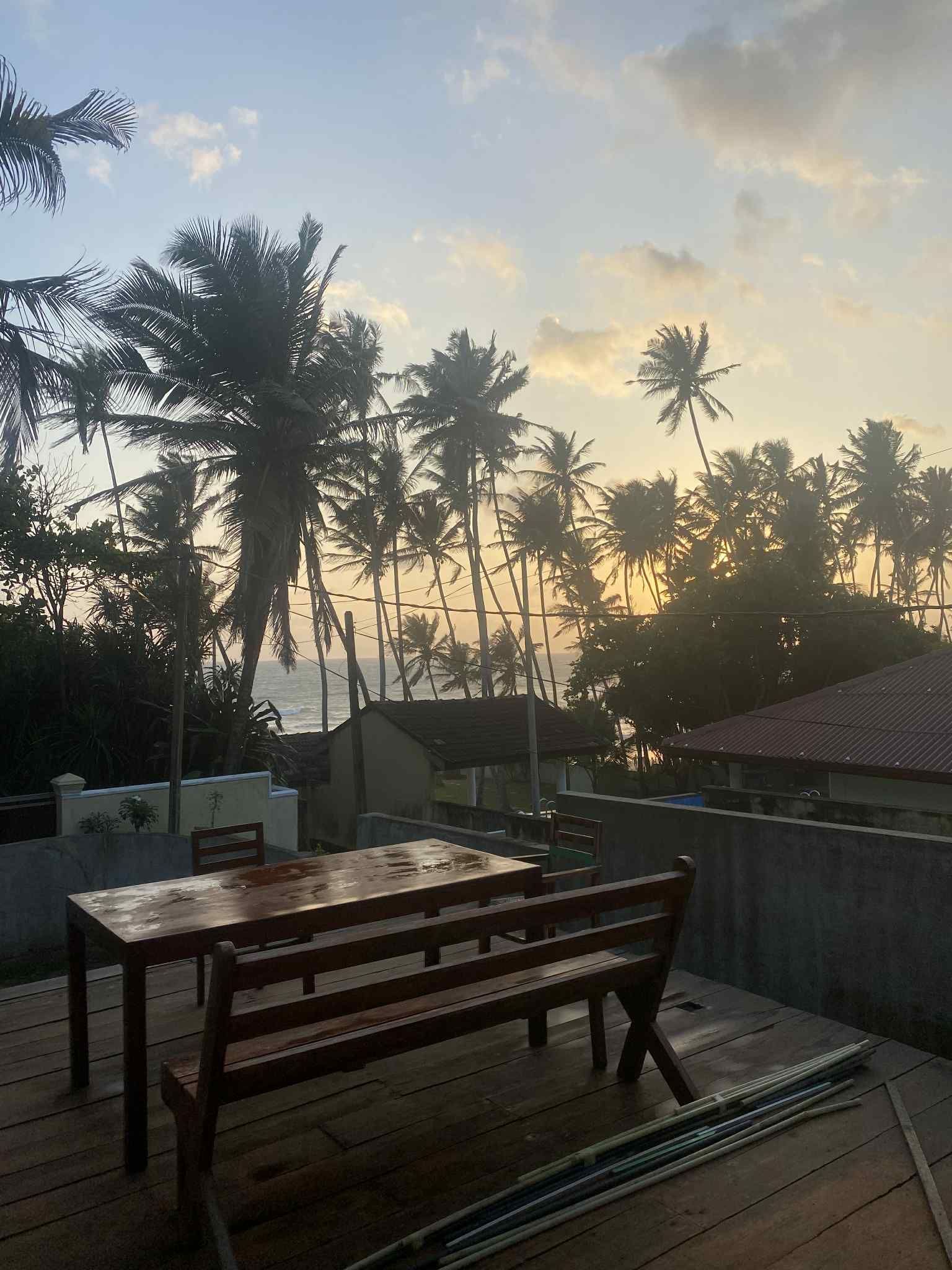 Wooden table and bench on a deck, with a view of palm trees, buildings, and a sunrise.