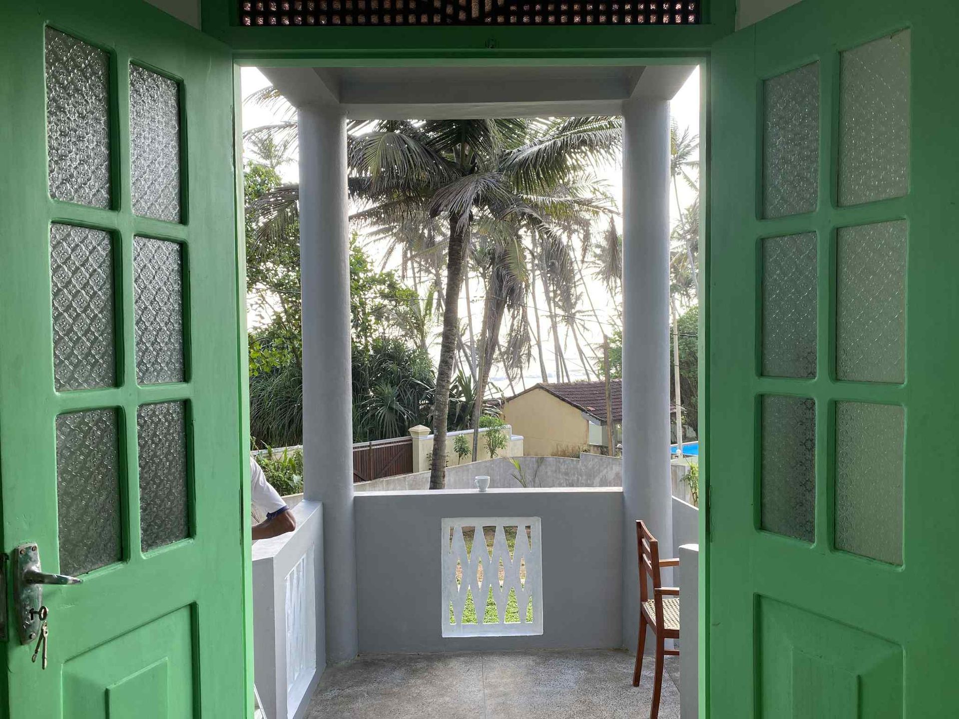 Green doors open to a balcony with palm trees and a small building in the distance.