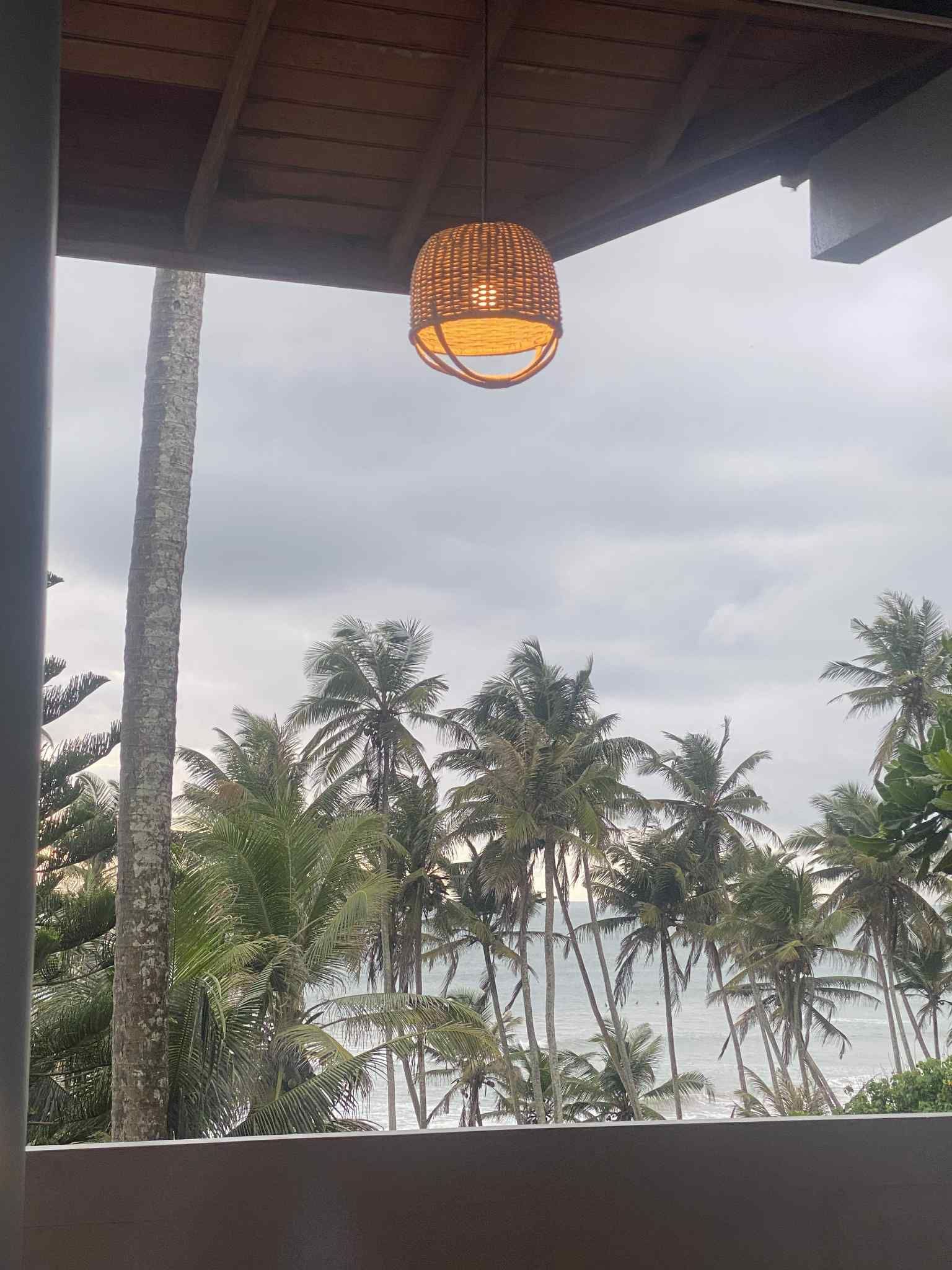 Wicker lamp hanging from a wood ceiling, overlooking a beach with palm trees and a cloudy sky.