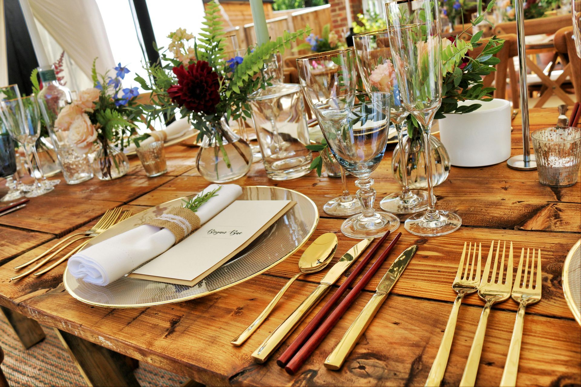 Wedding table setting: rustic wood table, gold flatware, floral centerpieces, crystal glasses, and menu.