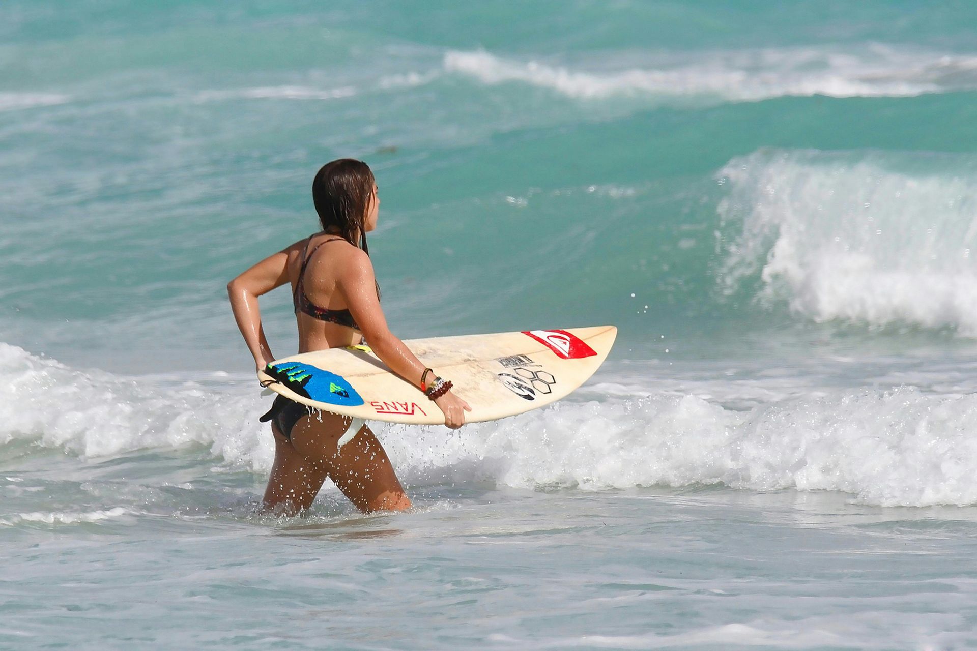 Woman in a bikini walks out of turquoise waves, carrying a surfboard.
