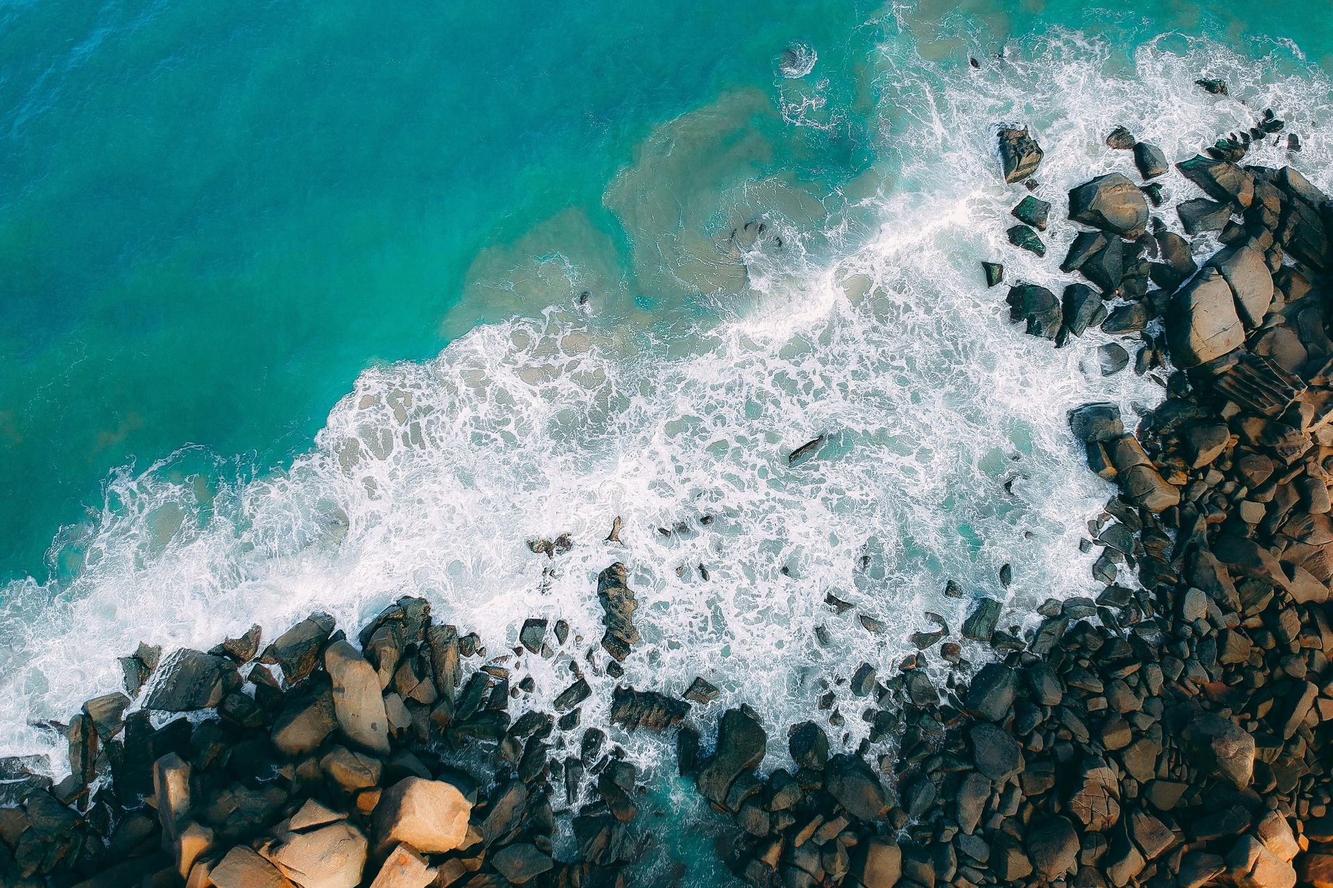 Ocean waves crashing against a rocky shoreline, turquoise water, overhead view.